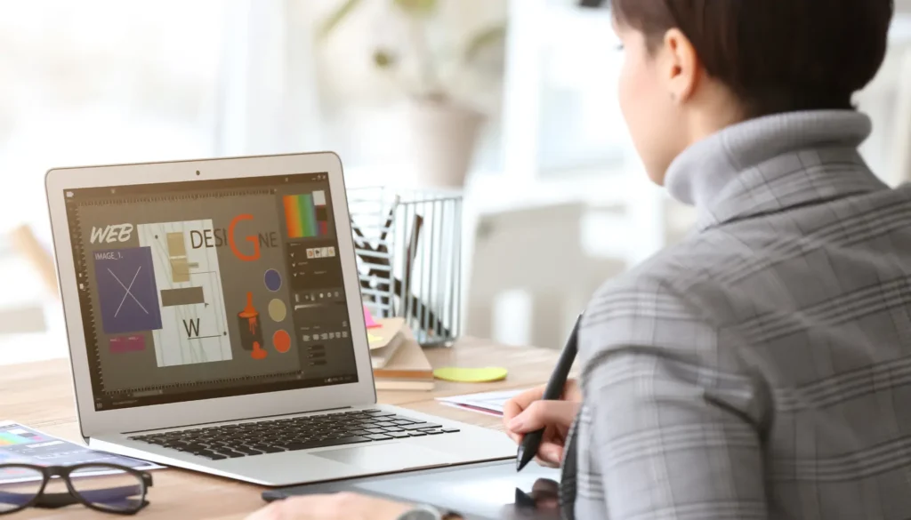 A designer works on a laptop, creating graphics, with colorful elements displayed on the screen and a creative workspace visible — reflecting the question, "Should Accessibility Overlay Tools Be Used as a Strategic Part of Your Accessibility Efforts?