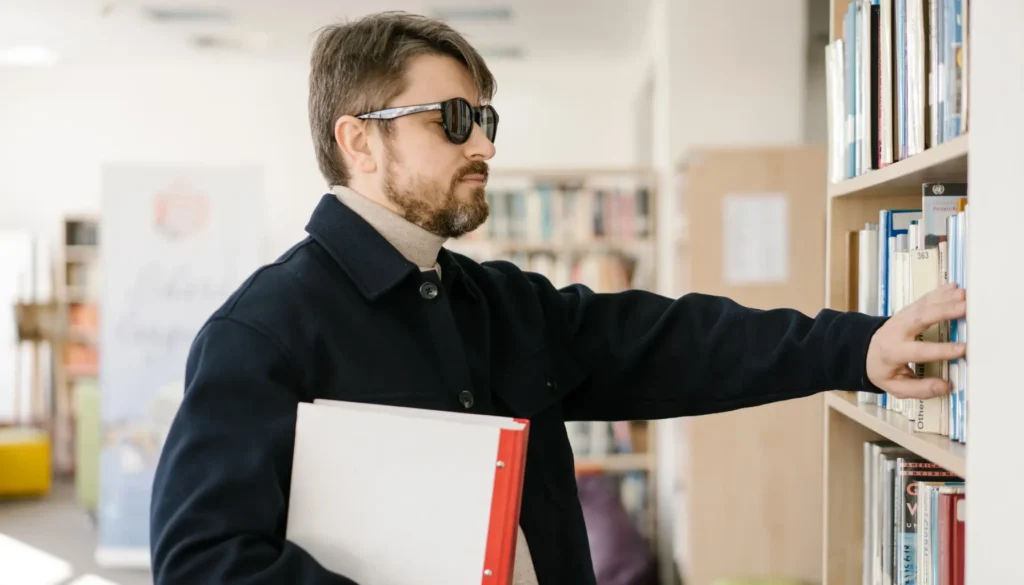 A person in a library, wearing a dark jacket, reaches for a book on a shelf while holding a binder in their other arm — illustrating how blind people’s brains rewire to adapt and enhance other senses.