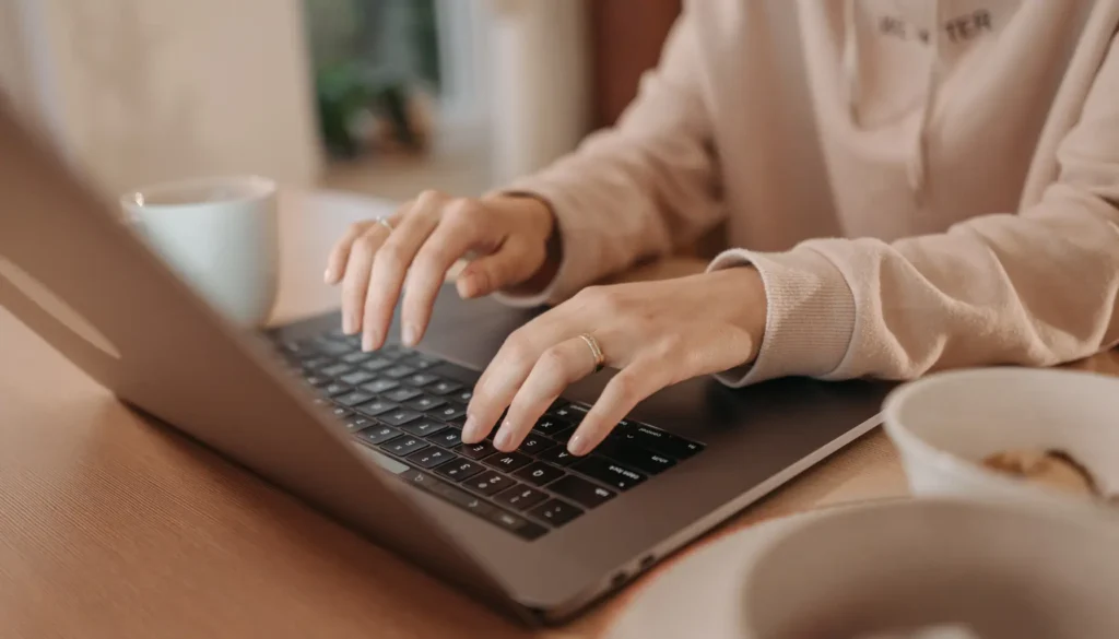 Hands typing on a laptop keyboard with a coffee cup and a bowl on a wooden table, representing work on Website Accessibility.