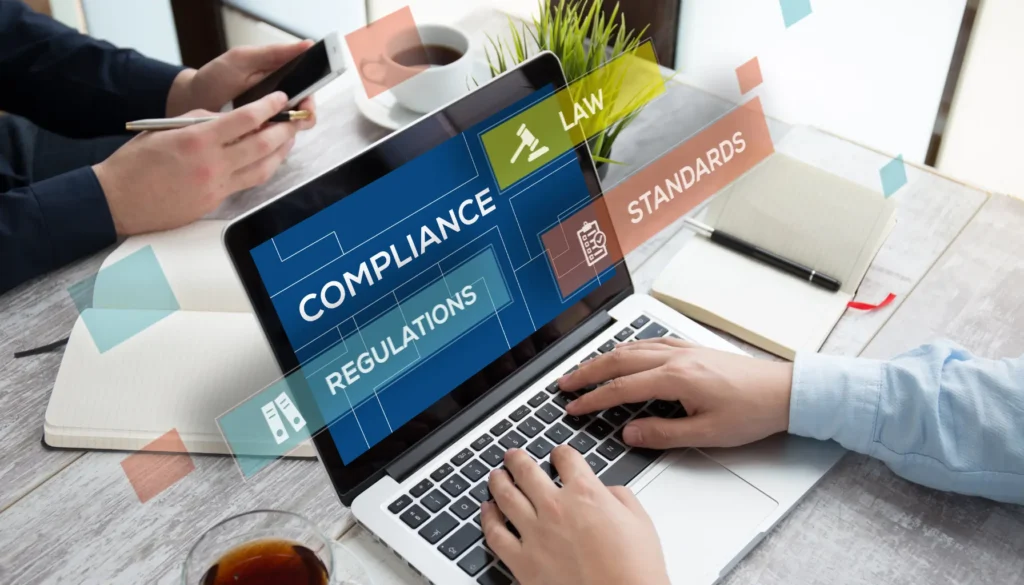 A person typing on a laptop displaying 'Compliance,' 'Regulations,' and 'Standards,' with a notebook and coffee in the background, illustrating AODA Compliance Requirements for Websites.