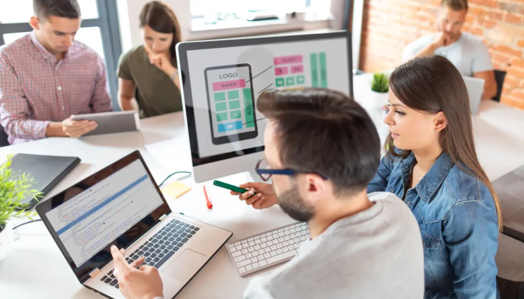 A group of people collaborates around a table, with laptops and tablets, discussing a mobile app design displayed on a computer screen, exploring the reasoning behind accessibility testing.