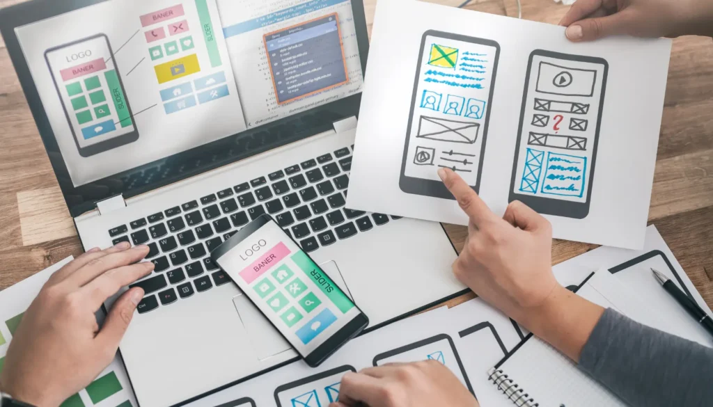 Two hands engage in a design discussion over smartphone mockups and a laptop displaying app layouts and coding screens, exploring how to test your website for accessibility.