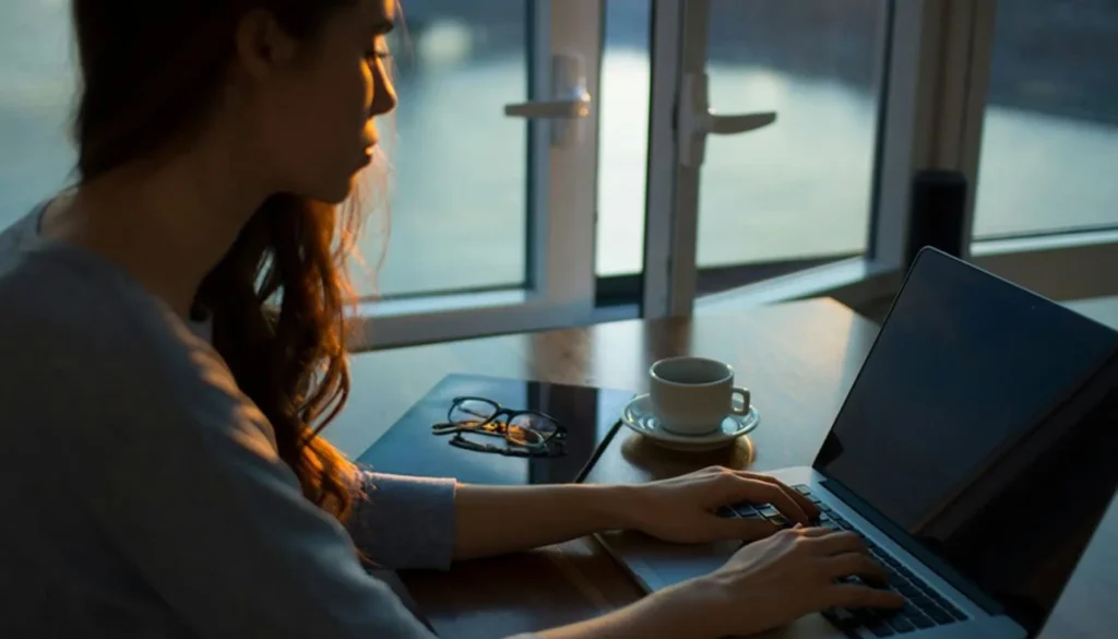 A woman sits at a wooden table, typing on a laptop, with a cup of coffee and glasses nearby, as sunlight streams through windows, highlighting the importance of the role of accessibility audits in improving user experience.