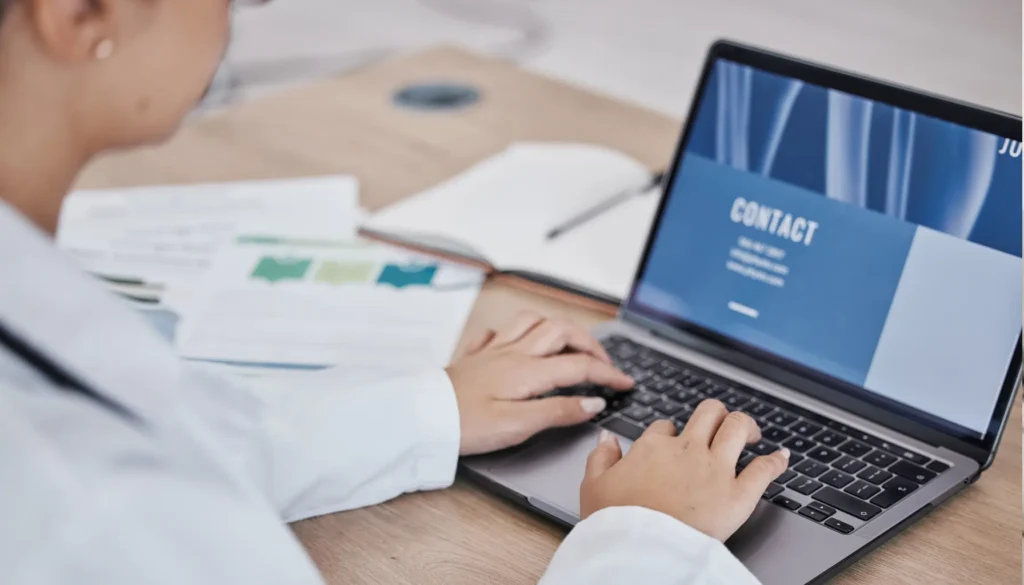 Professional in a white lab coat typing on a laptop displaying a contact page, with documents scattered on a wooden table — highlighting Document Remediation in Accessibility.