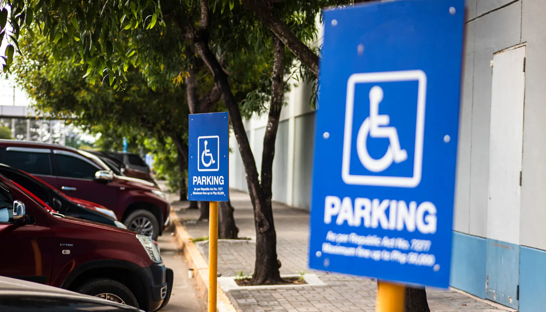 Two blue parking signs for disabled parking, surrounded by trees and parked cars, indicating reserved spaces in a lot, meeting accessible parking requirements.